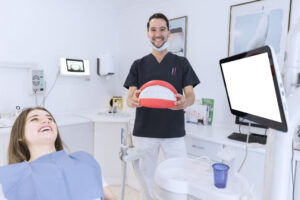 Dentist holding teeth model while patient relaxes in dental chair.